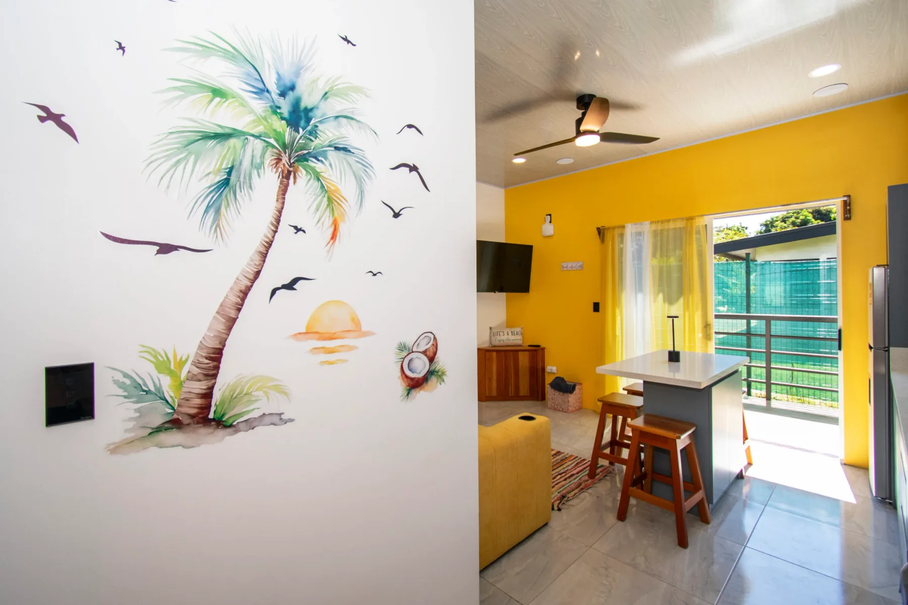 A bright yellow living area opens onto a balcony, with a kitchen island and stools.