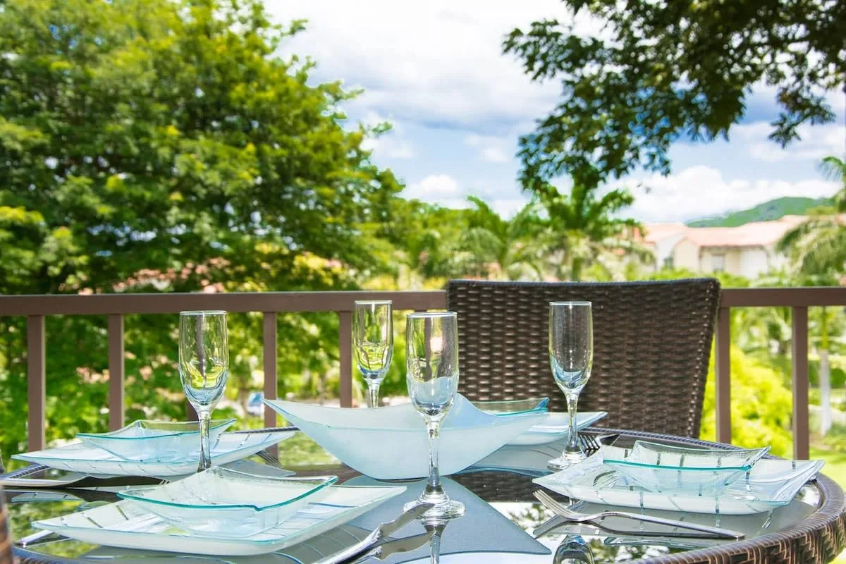 An outdoor dining table is set with glassware and plates beside a leafy view.