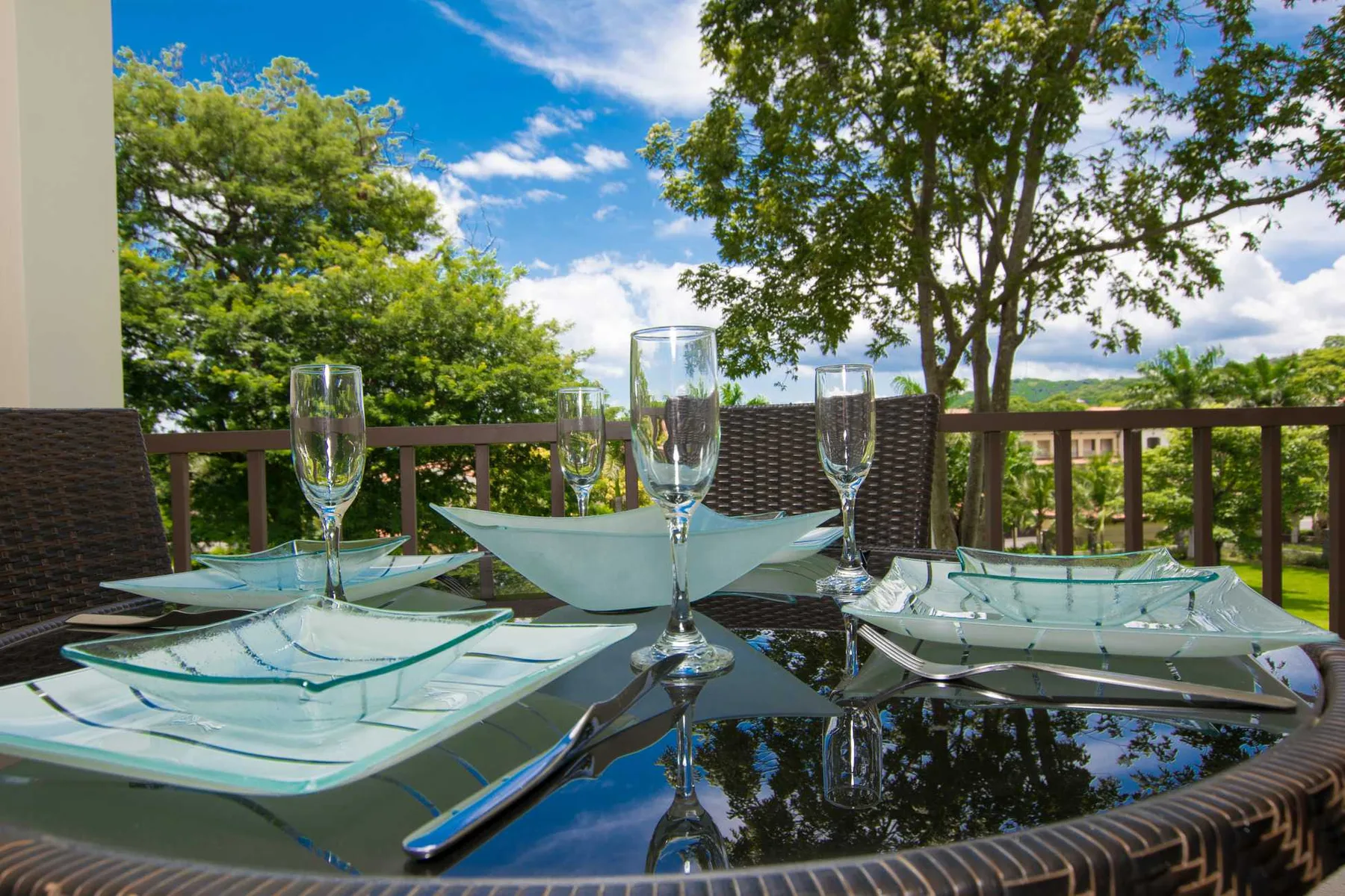 An outdoor dining table is set with glassware beside a wooden deck railing.