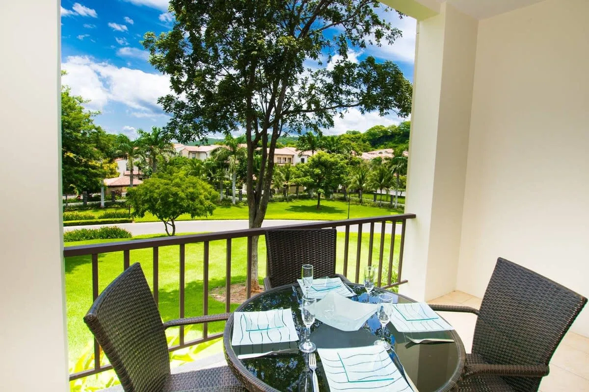A balcony dining area overlooks a green lawn and trees.