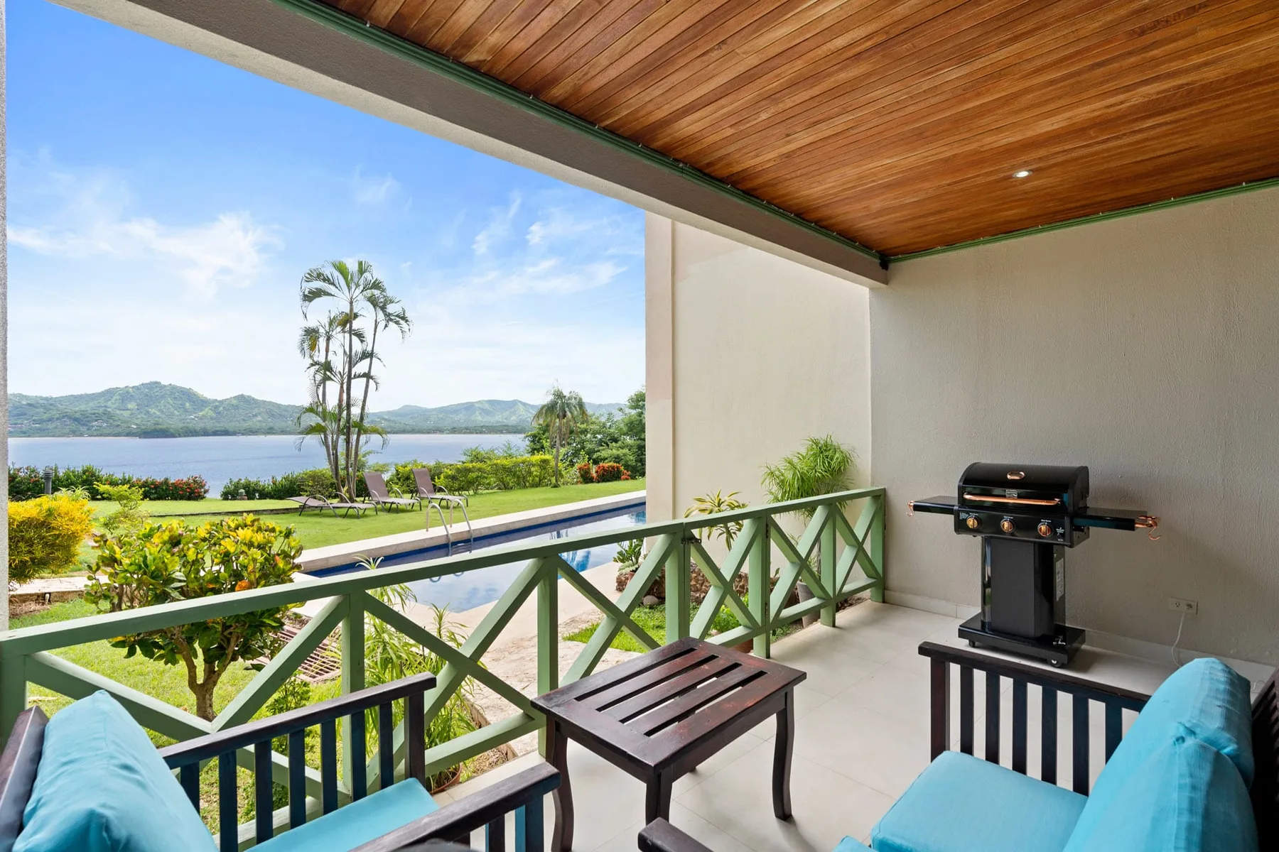 A covered balcony has seating and a grill, with a view of the garden and water.