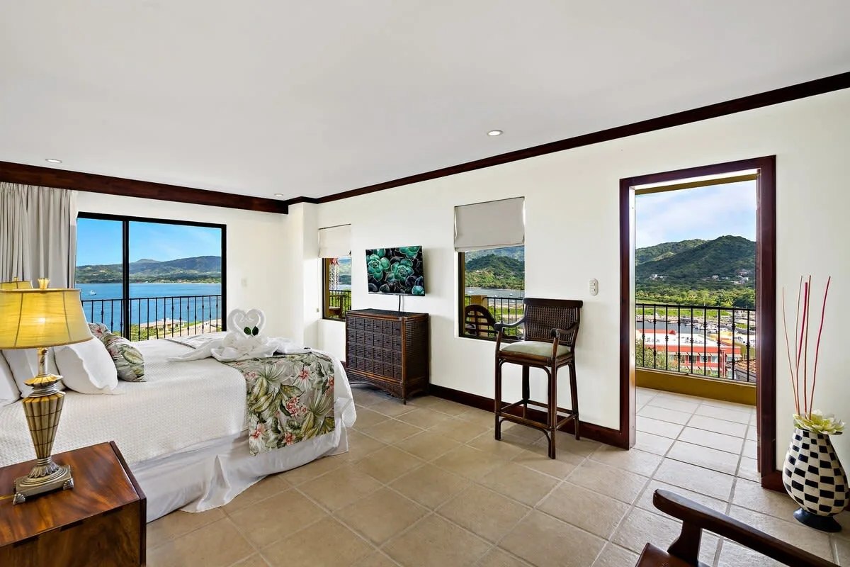 Un dormitorio luminoso con cama, asientos y vistas a través de grandes ventanas.