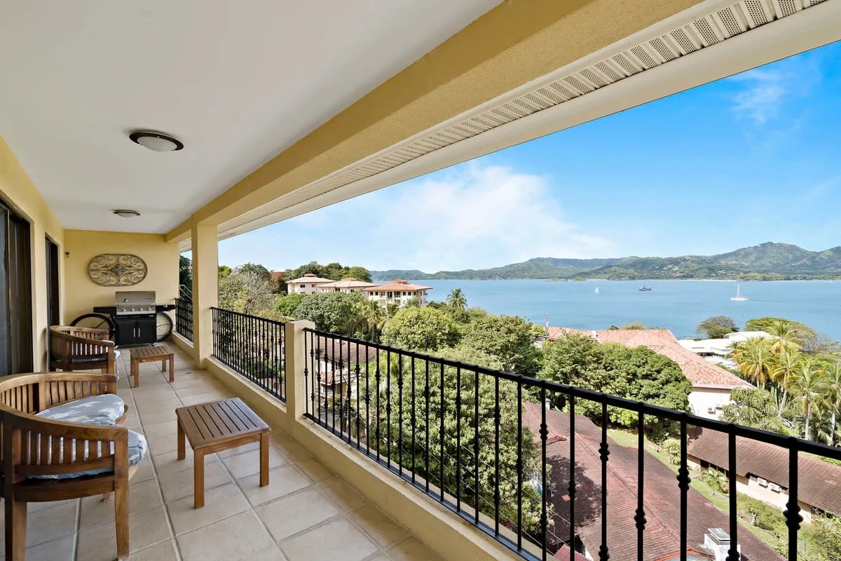 A covered balcony with seating overlooks a bay and green hills.