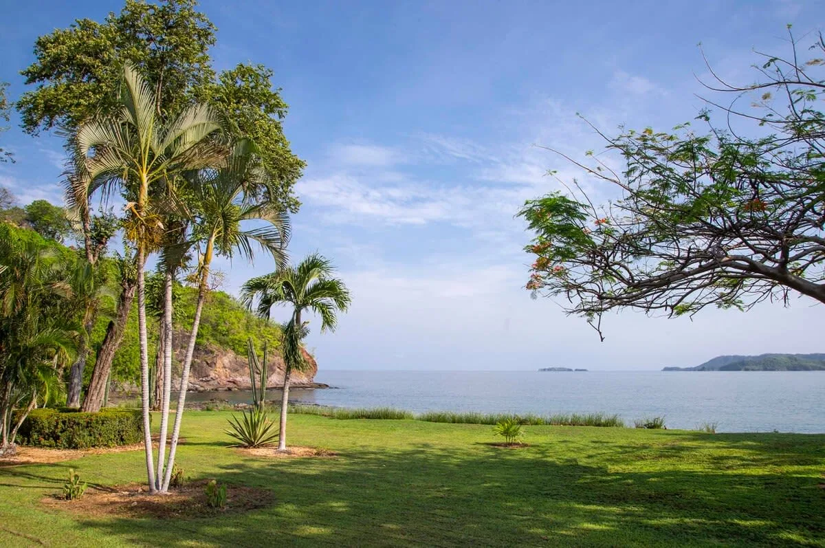 A grassy tropical waterfront garden overlooks calm water beneath a partly cloudy sky.