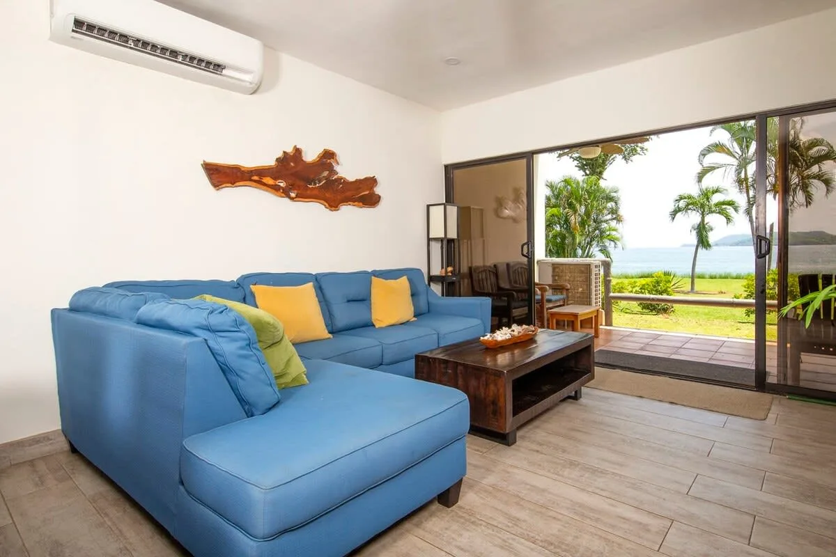 A bright living room with blue seating and a wide window overlooking tropical greenery.