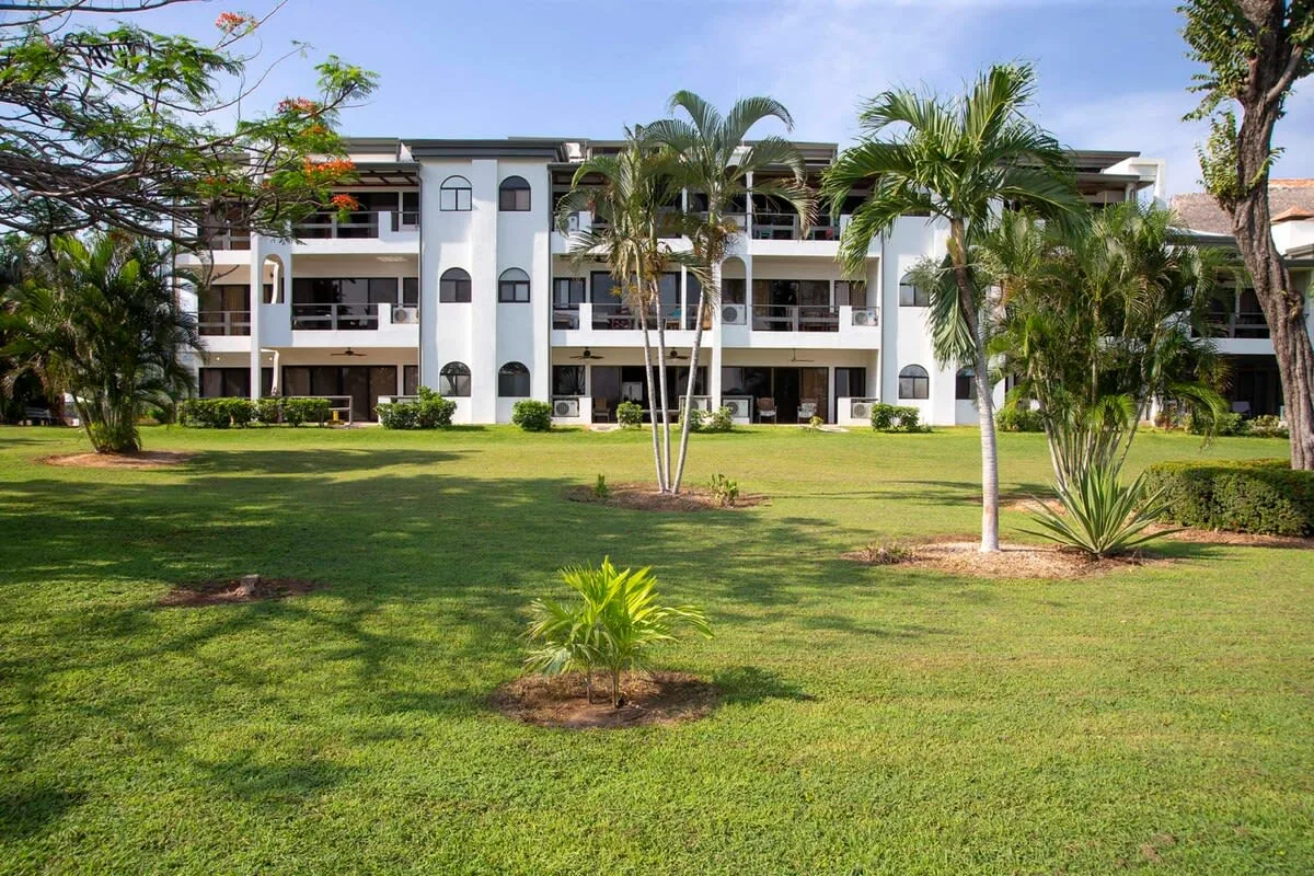 A multi-story building stands beyond a landscaped lawn with palm trees.