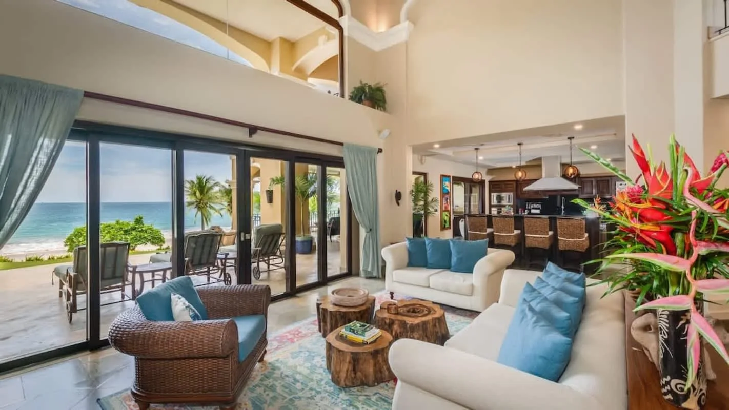 A bright living room opens onto a patio with ocean views and tropical plants.