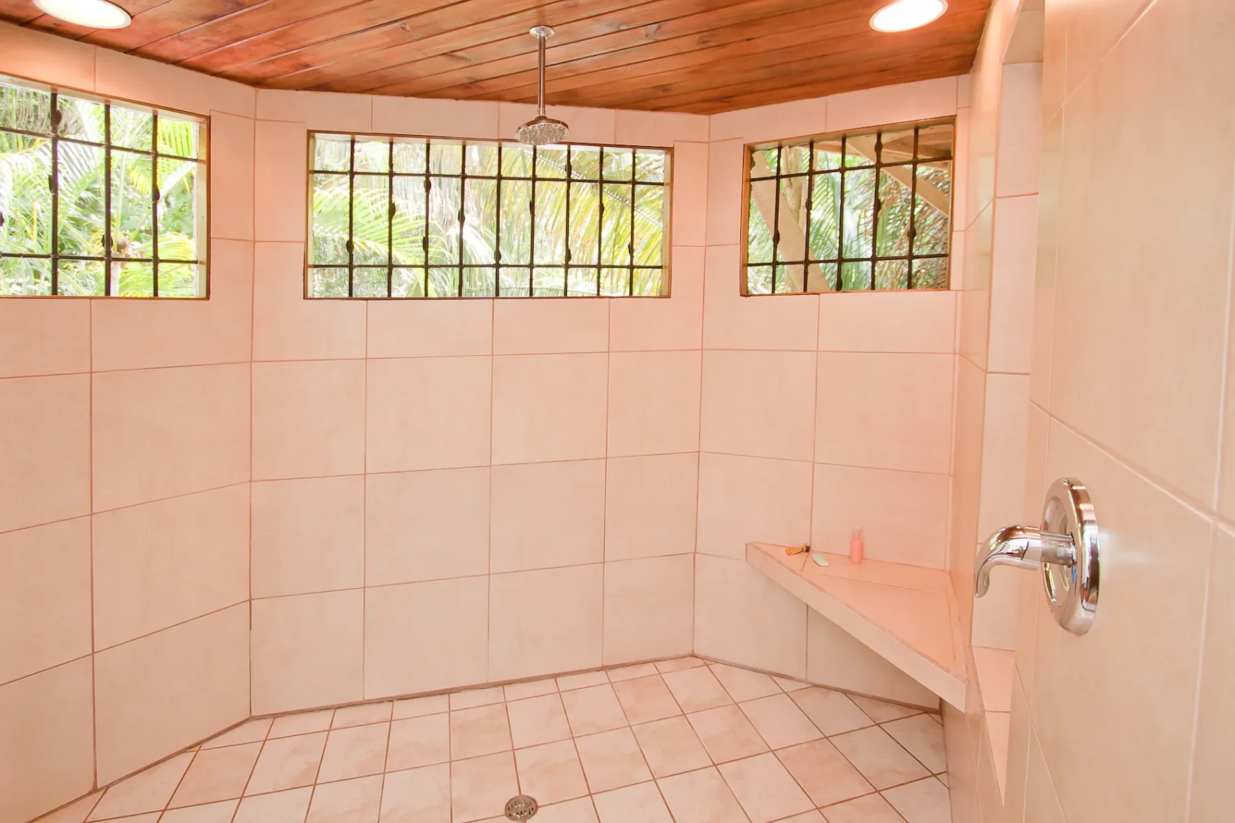 Bali Tree House · 24. A tiled shower room features a wooden ceiling, ceiling-mounted shower head, built-in bench, and multiple barred windows overlooking lush foliage.