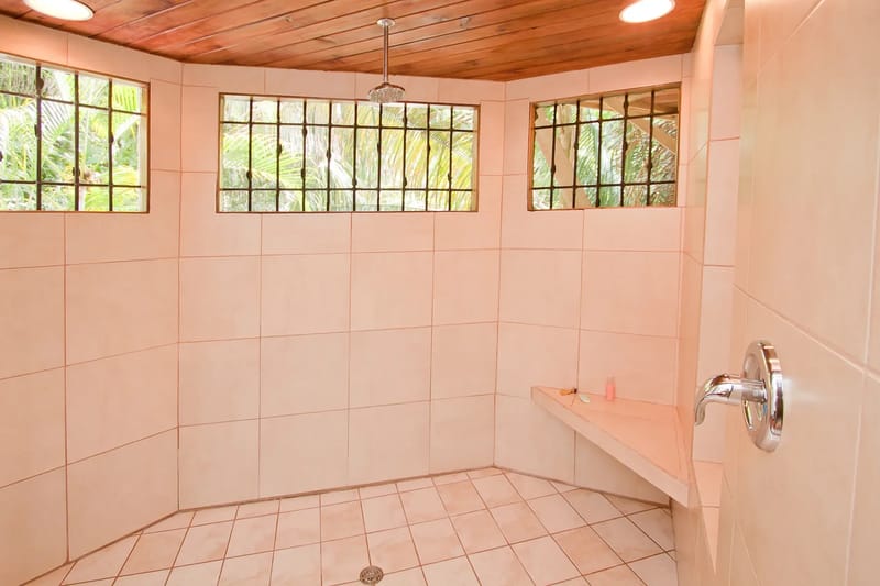 Bali Tree House · 24. A tiled shower room features a wooden ceiling, ceiling-mounted shower head, built-in bench, and multiple barred windows overlooking lush foliage.