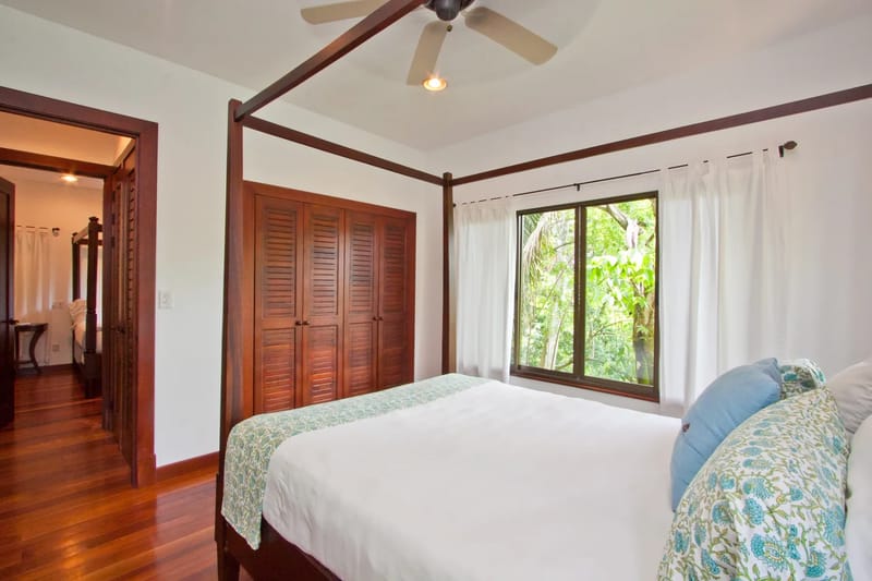 Bali Tree House · 26. A bedroom with a four-poster bed, a louvered wooden closet, a ceiling fan, and a window overlooking dense foliage.