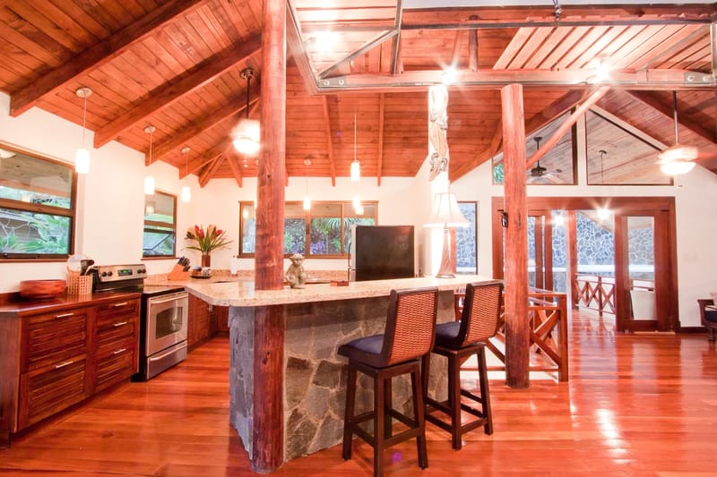 Bali Tree House · 16. A kitchen area features a stainless steel oven, dark wood cabinetry, granite countertops, and an island with two bar stools, set within a room with wooden vaulted ceilings, wood floors, and large windows.