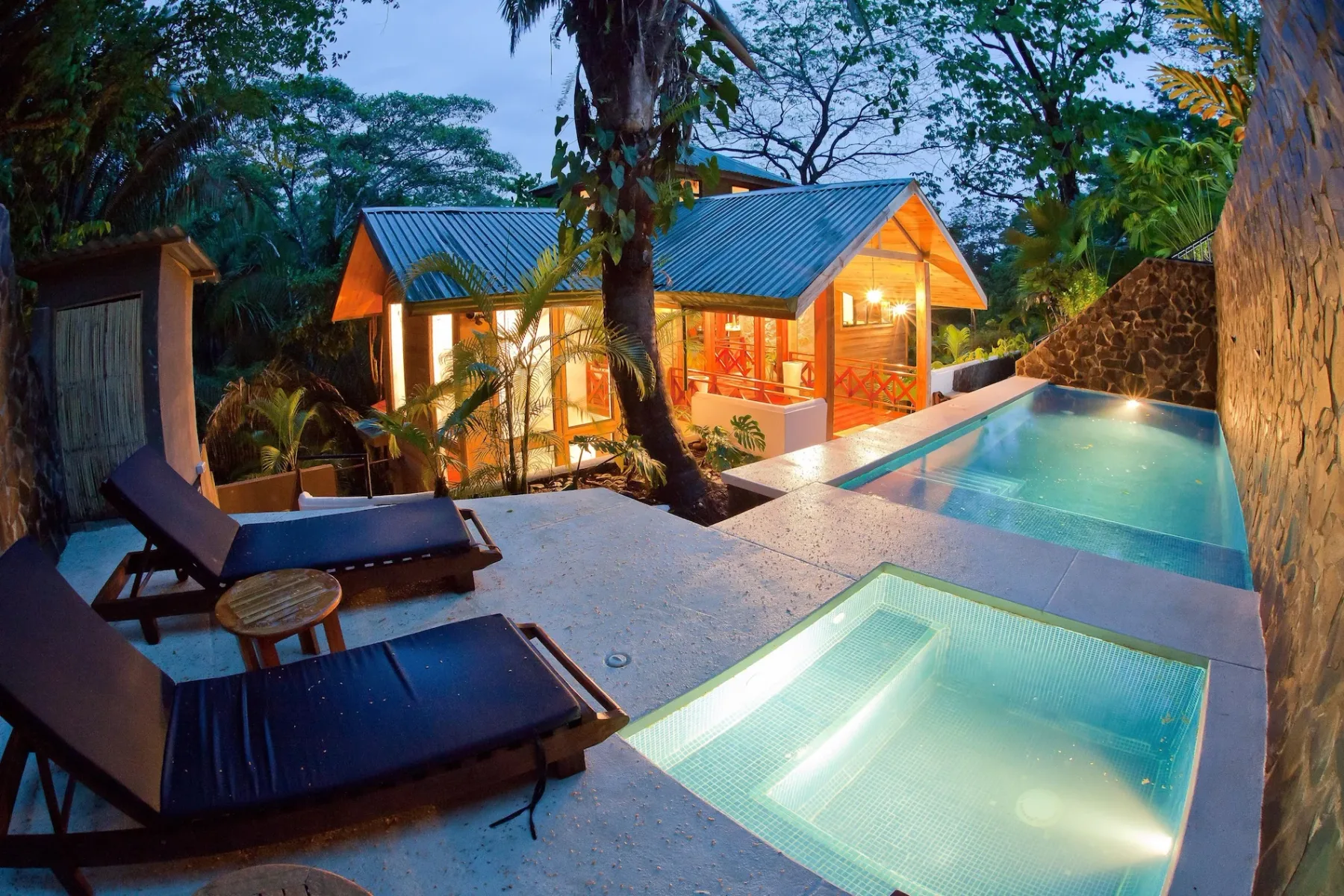 Bali Tree House · 8. An illuminated private plunge pool and separate jacuzzi sit on a stone terrace overlooking a warm-lit wooden villa and lush jungle foliage at twilight.