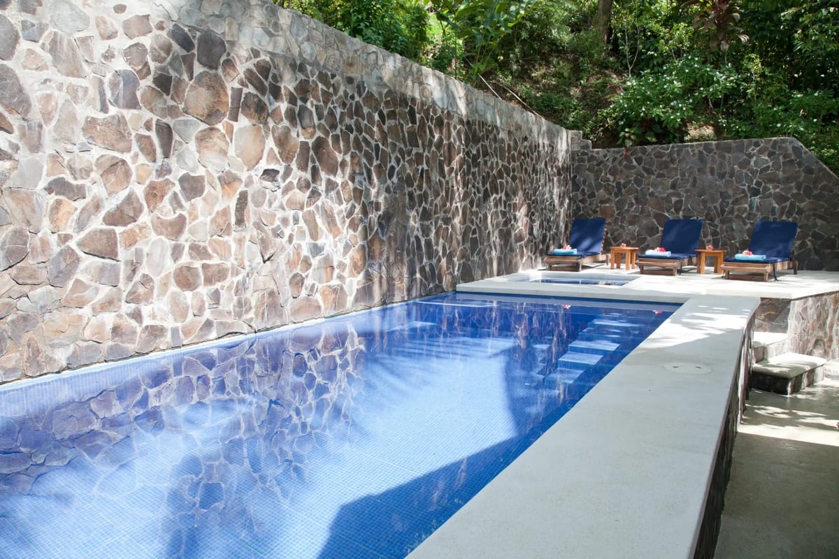 Bali Tree House · 5. An outdoor tiled swimming pool with an integrated spa is situated next to a stone wall, with lounge chairs on a concrete deck.