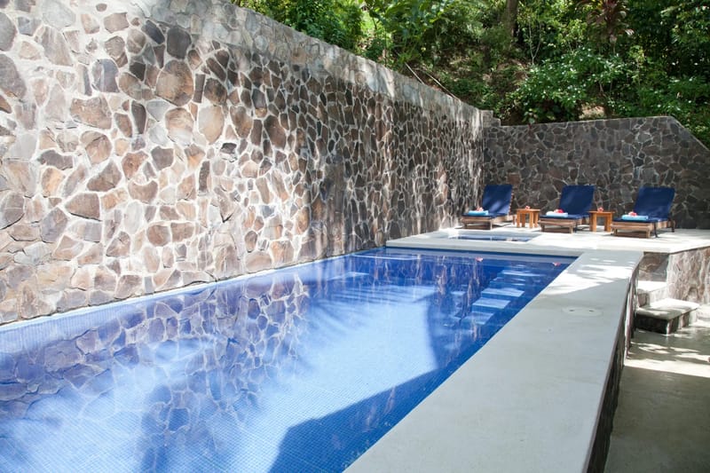 Bali Tree House · 5. An outdoor tiled swimming pool with an integrated spa is situated next to a stone wall, with lounge chairs on a concrete deck.