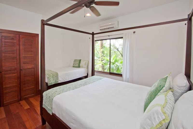 Bali Tree House · 27. A bedroom features a wooden four-poster bed, a twin bed, louvered closet doors, wood flooring, a window overlooking trees, a ceiling fan, and an air conditioning unit.