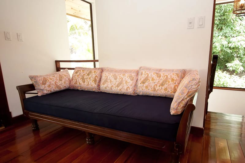 Bali Tree House · 19. A wooden daybed with a dark cushion and patterned pillows is situated on a polished wood floor between white walls and windows.