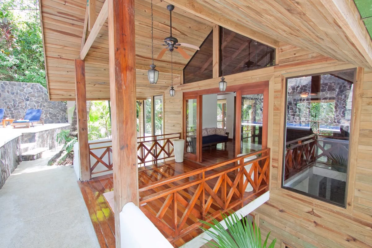 Bali Tree House · 4. A wooden terrace with a railing and ceiling fans leads to an outdoor patio with lounge chairs, surrounded by lush tropical greenery, with a view into an interior room.
