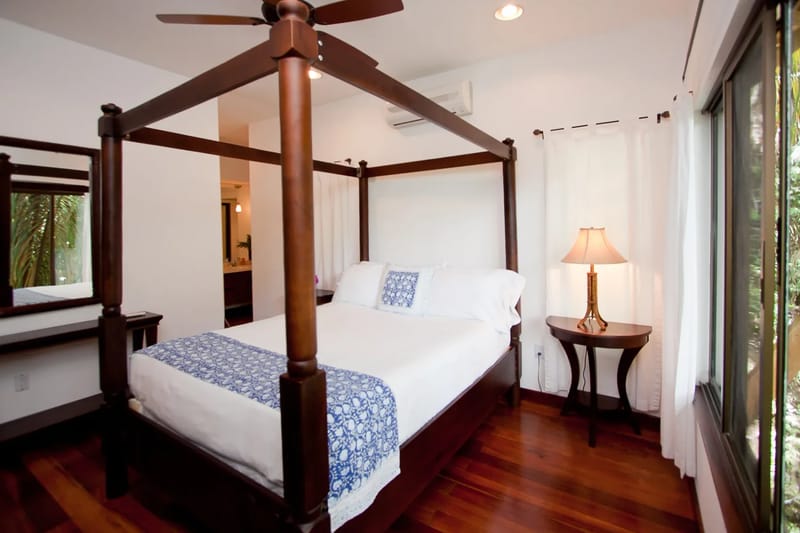 Bali Tree House · 22. A bedroom features a dark wood four-poster bed with white and blue bedding, a bedside table with a lamp, a ceiling fan, hardwood floors, and a large window.