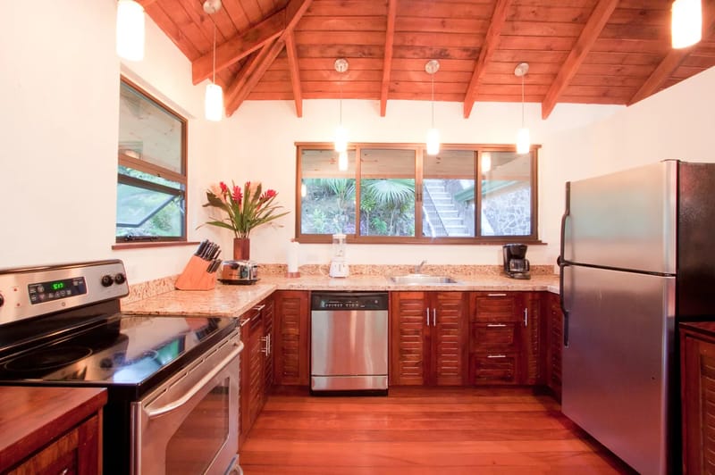 Bali Tree House · 17. A well-appointed kitchen features rich wooden floors, cabinetry, and a vaulted ceiling, complemented by stainless steel appliances and windows overlooking the tropical jungle.