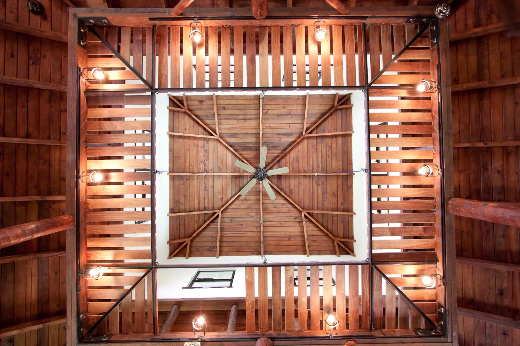 Bali Tree House · 15. A wooden ceiling structure with exposed beams, slatted sections, a central ceiling fan, and multiple mounted light fixtures.