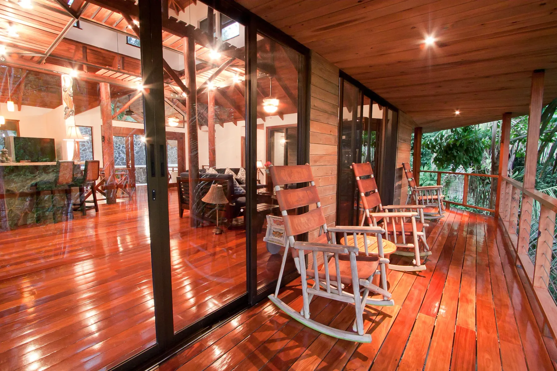 Bali Tree House · 14. A wooden terrace with rocking chairs overlooks lush foliage, with a living room visible through large glass doors.