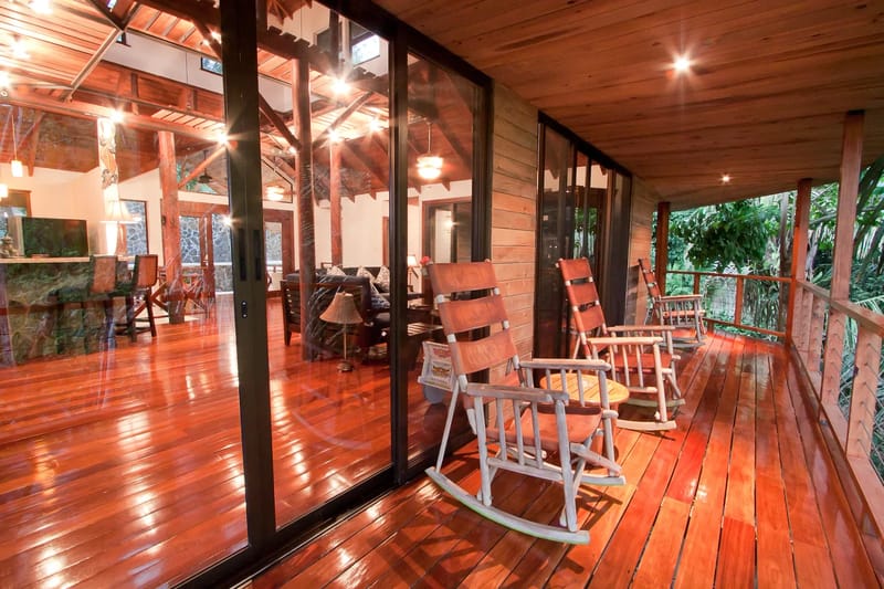 Bali Tree House · 14. A wooden terrace with rocking chairs overlooks lush foliage, with a living room visible through large glass doors.