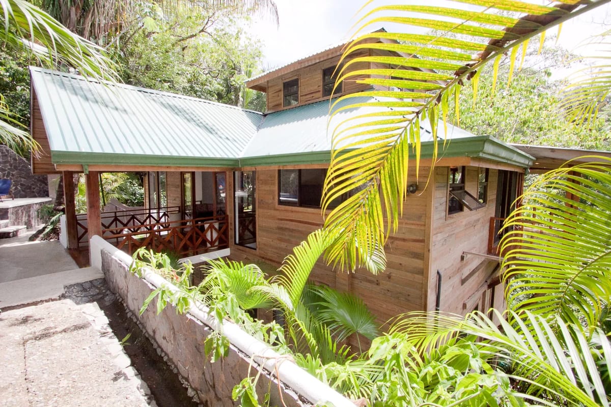 Bali Tree House · 3. An exterior view of a wooden house with a green metal roof and covered balcony, surrounded by tropical foliage.