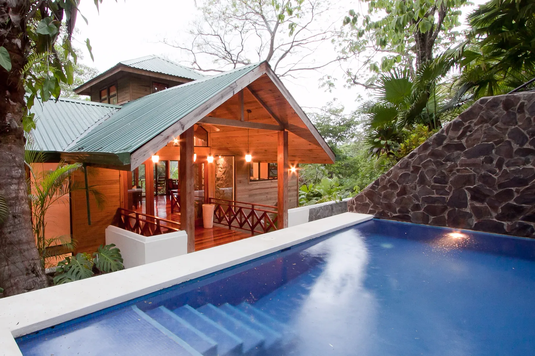 Bali Tree House · 10. A wooden house with a green metal roof features an illuminated porch and an adjacent outdoor swimming pool, surrounded by lush vegetation.