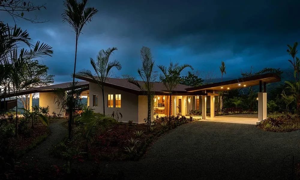 Casa Grande del Mar · 6. The modern villa, with its angled rooflines and warmly lit interior, is nestled amidst lush tropical landscaping and palm trees, featuring a covered entry and carport at twilight.