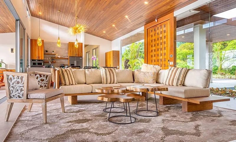 Casa Grande del Mar · 1. An open-concept living room with a sloped wood-paneled ceiling and a large sectional sofa connects to an adjacent kitchen, with full-height glass walls offering views of the surrounding jungle.