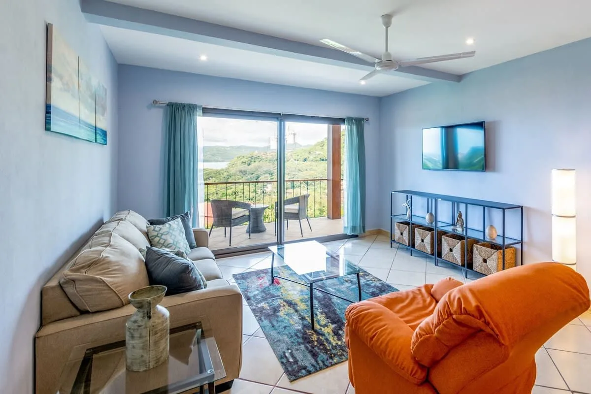 Una sala de estar con asientos, un televisor y puertas corredizas de vidrio hacia un balcón.