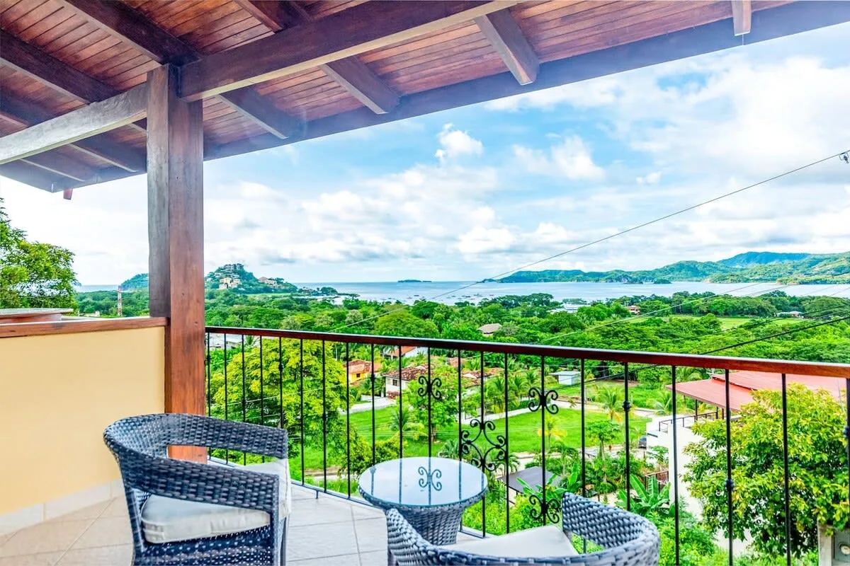 A covered terrace overlooks green hills and a bay beyond the railing.