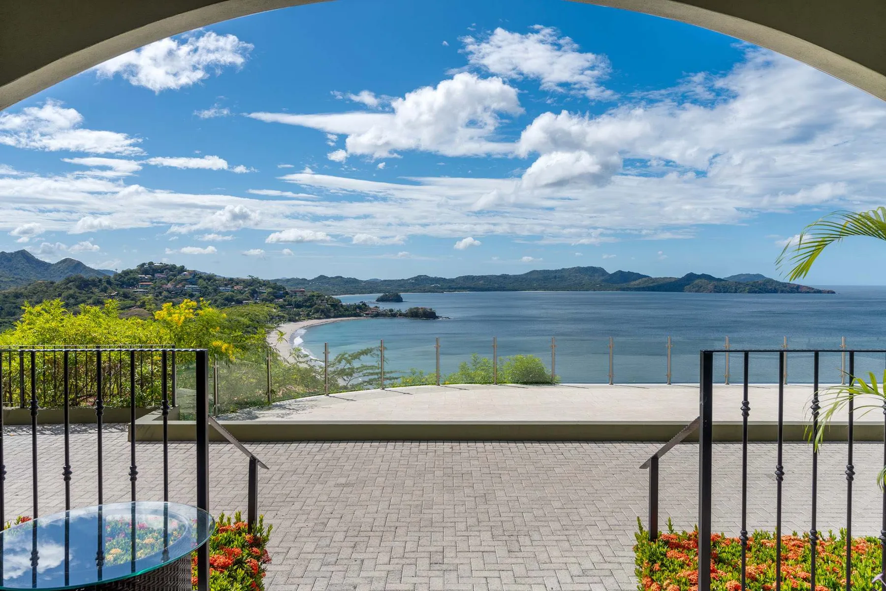 A paved terrace overlooks a broad bay and green hills beneath a partly cloudy sky.