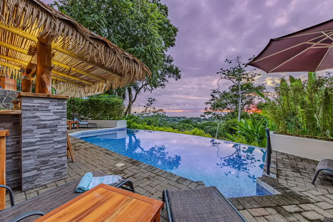An outdoor pool overlooks lush greenery and the ocean beneath a cloudy sky.