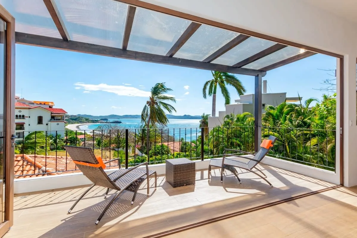 A covered terrace with lounge seating overlooks palm trees and the water.