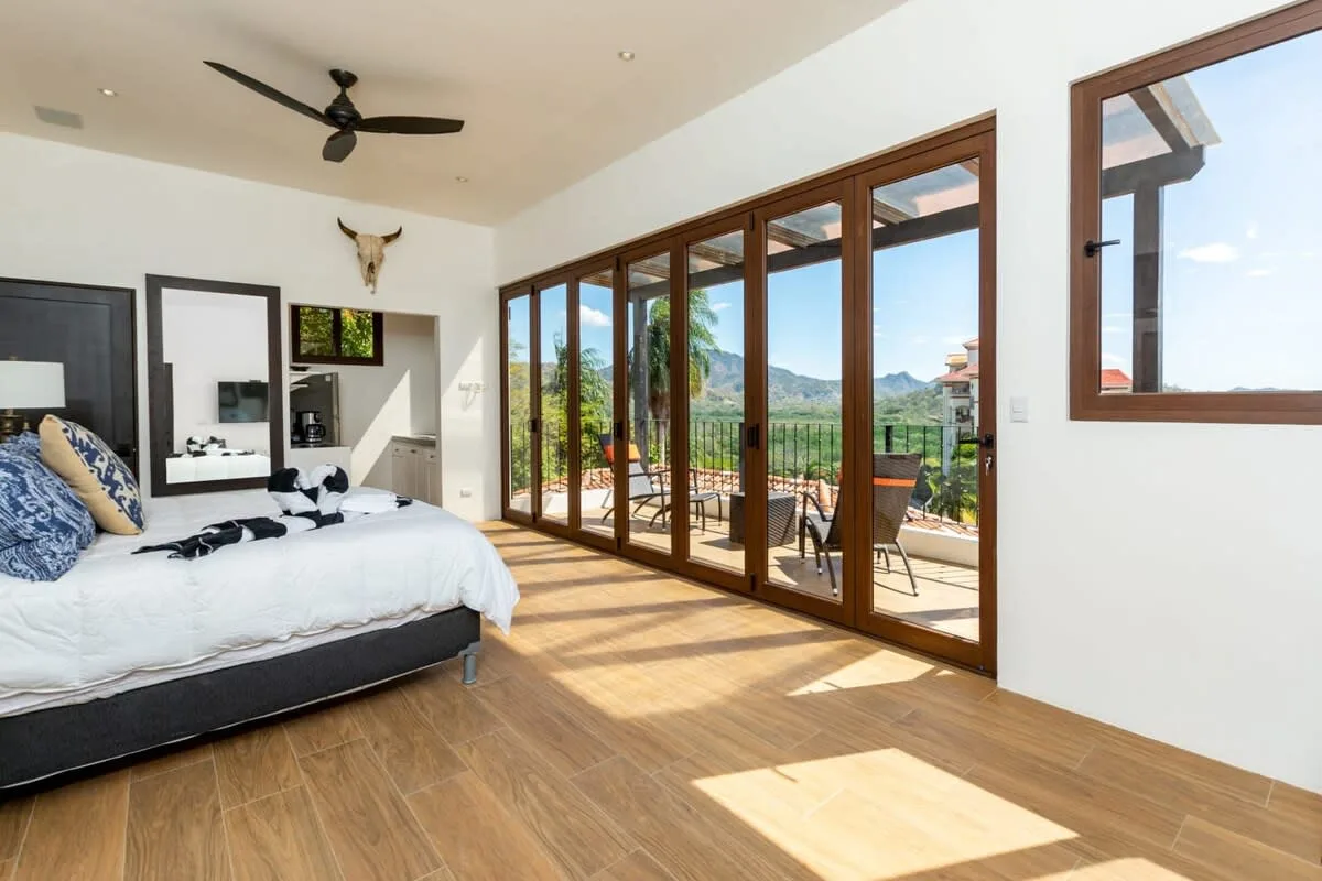 A bright bedroom opens onto a sunlit terrace through large glass doors.