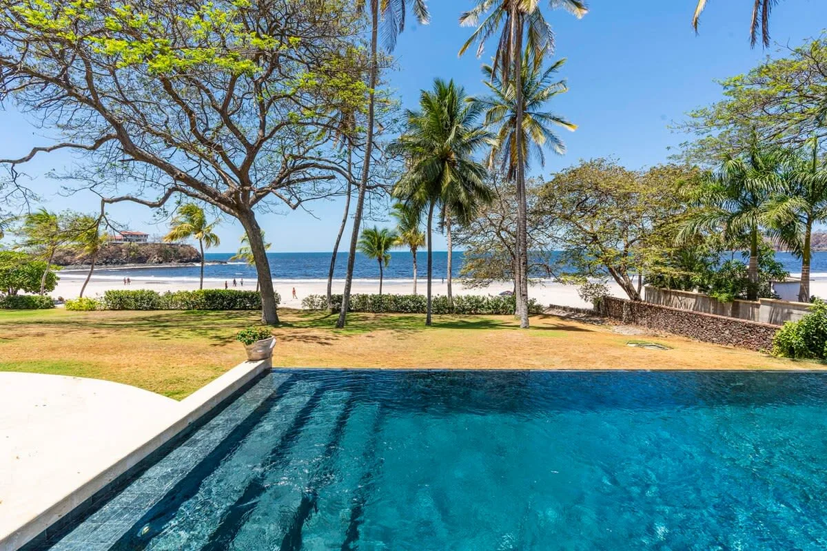 A swimming pool overlooks a sandy beach and palm-lined shoreline.