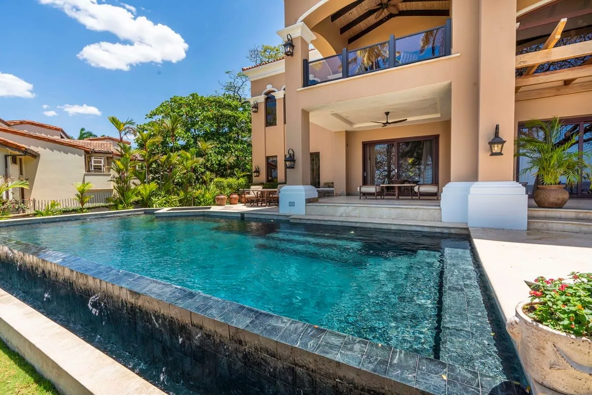 A swimming pool sits beside a covered patio and a multi-story building.