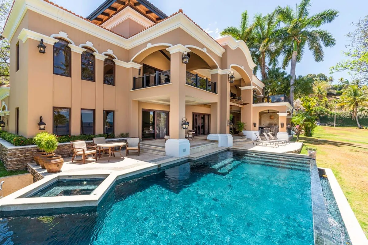 A large house overlooks a swimming pool and patio seating.