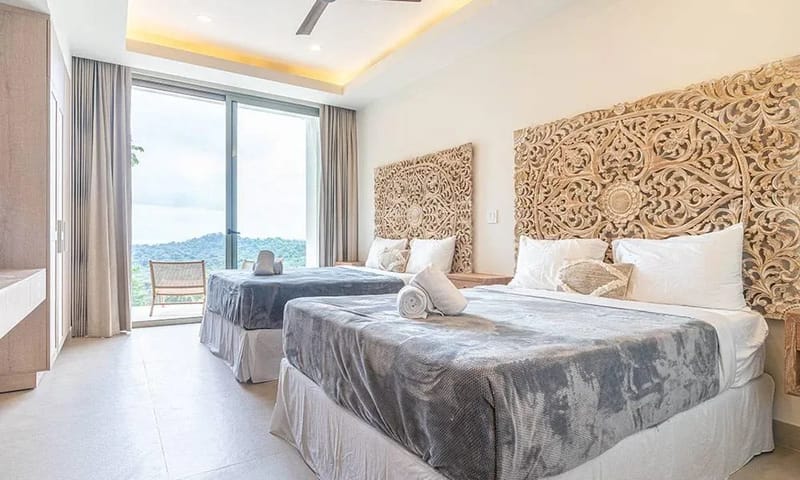 Casa Frenchie & Guest House · 25. This bright bedroom features two queen beds with intricately carved headboards, receiving ample natural light from large sliding doors that open to a jungle and distant ocean view.