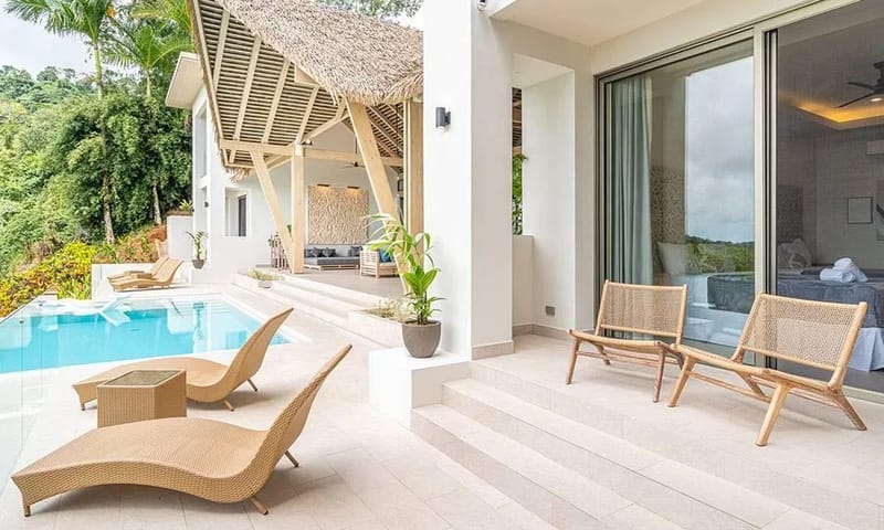 Casa Frenchie & Guest House · 26. A bright outdoor terrace with an infinity pool and lounge chairs connects to a bedroom through sliding glass doors, while a thatched-roof open-air lounge overlooks the jungle-covered hills.