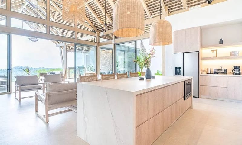 Casa Frenchie & Guest House · 9. This open-plan kitchen features a large island, light wood cabinetry, and vaulted ceilings with exposed beams, receiving abundant natural light from sliding glass doors that offer distant jungle views.