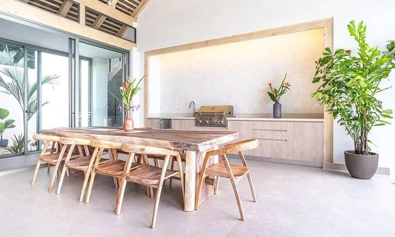 Casa Frenchie & Guest House · 14. This open-air dining area features a live-edge wooden table and chairs, an integrated outdoor kitchen with a grill, and vaulted ceilings, all bathed in natural light and connected to tropical greenery through large sliding glass doors.