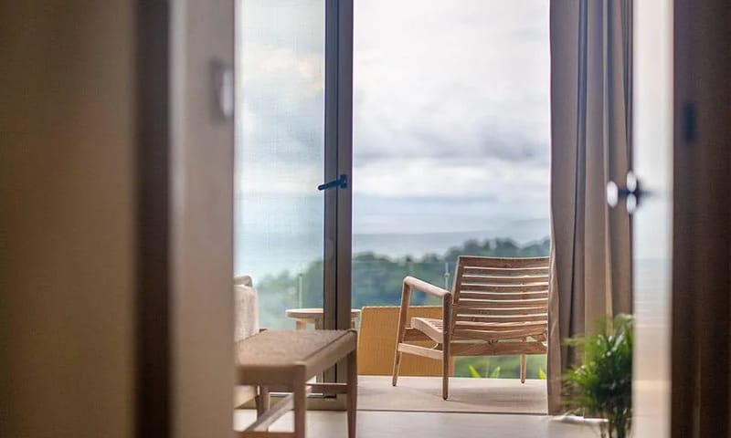 Casa Frenchie & Guest House · 27. A bright bedroom opens through wide glass doors to a private terrace, revealing views of the surrounding jungle and distant ocean.