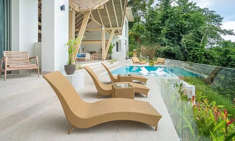 Casa Frenchie & Guest House · 28. An expansive outdoor pool terrace with multiple loungers and an infinity pool offers lush jungle views from behind a clear glass railing, connecting to an open-air living area under a vaulted thatched roof.