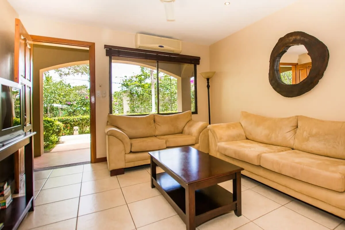 A living room with beige seating, a coffee table, and a garden view through the open doorway.