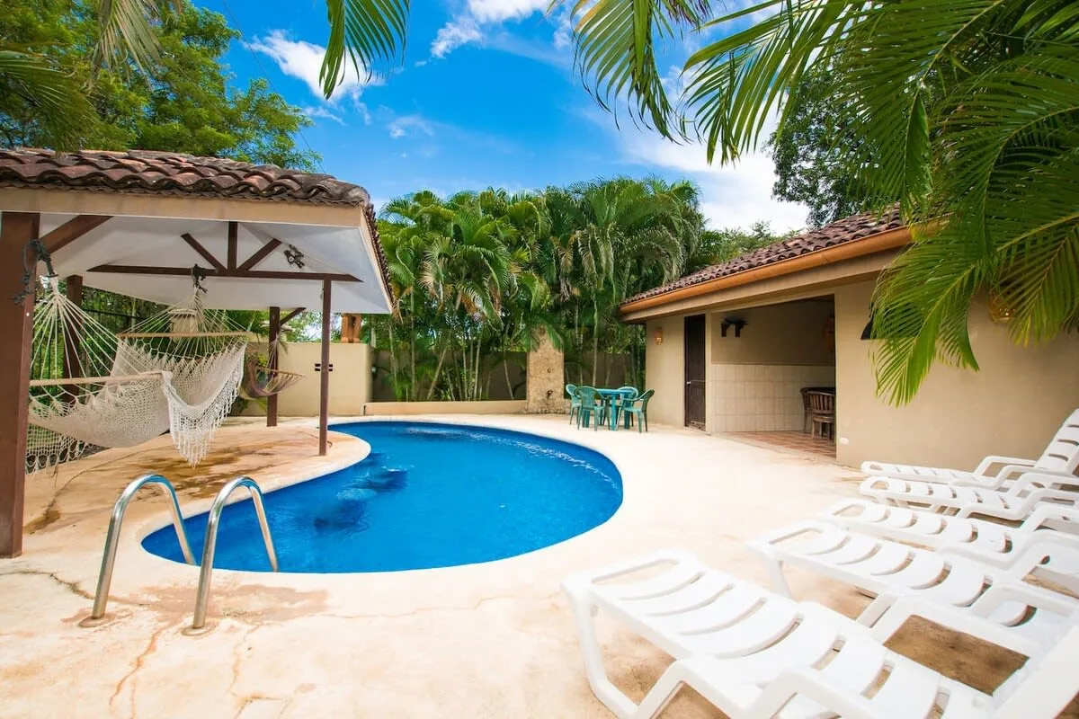 A tropical courtyard pool is surrounded by lounge chairs, palms, and a shaded hammock area.