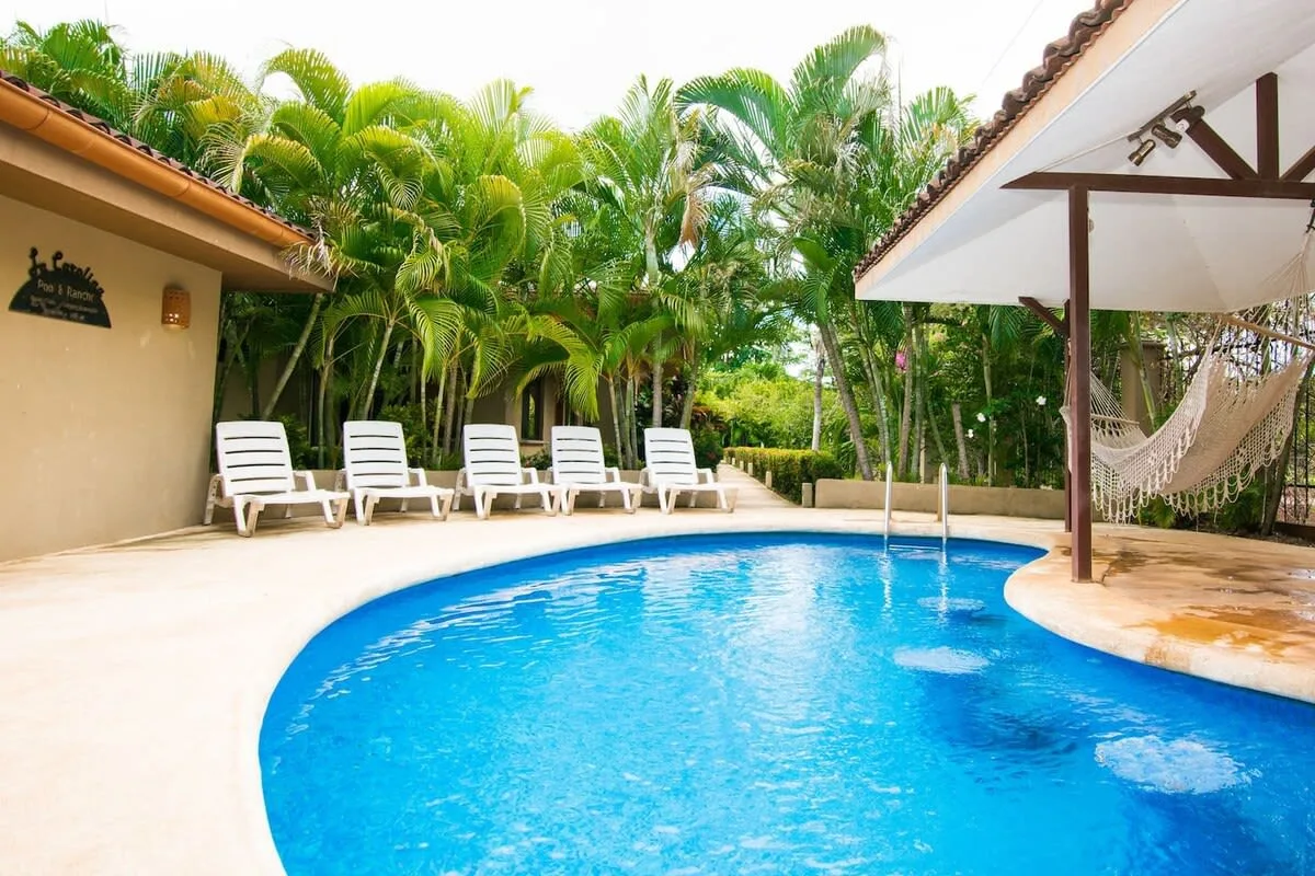 An outdoor swimming pool beside a shaded patio and tropical greenery.