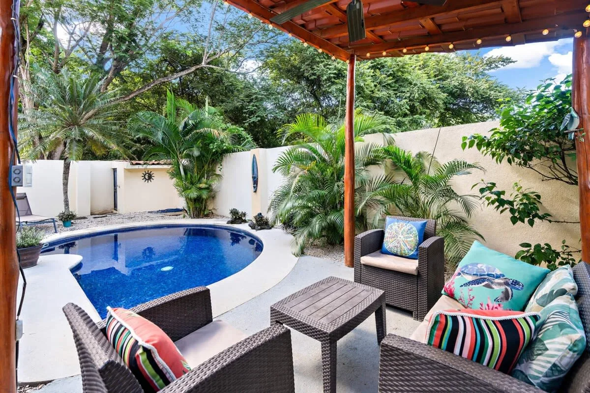 A covered outdoor patio with seating beside a small pool and tropical plants.