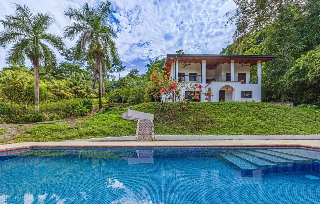 A swimming pool sits in front of a two-story house amid tropical greenery.